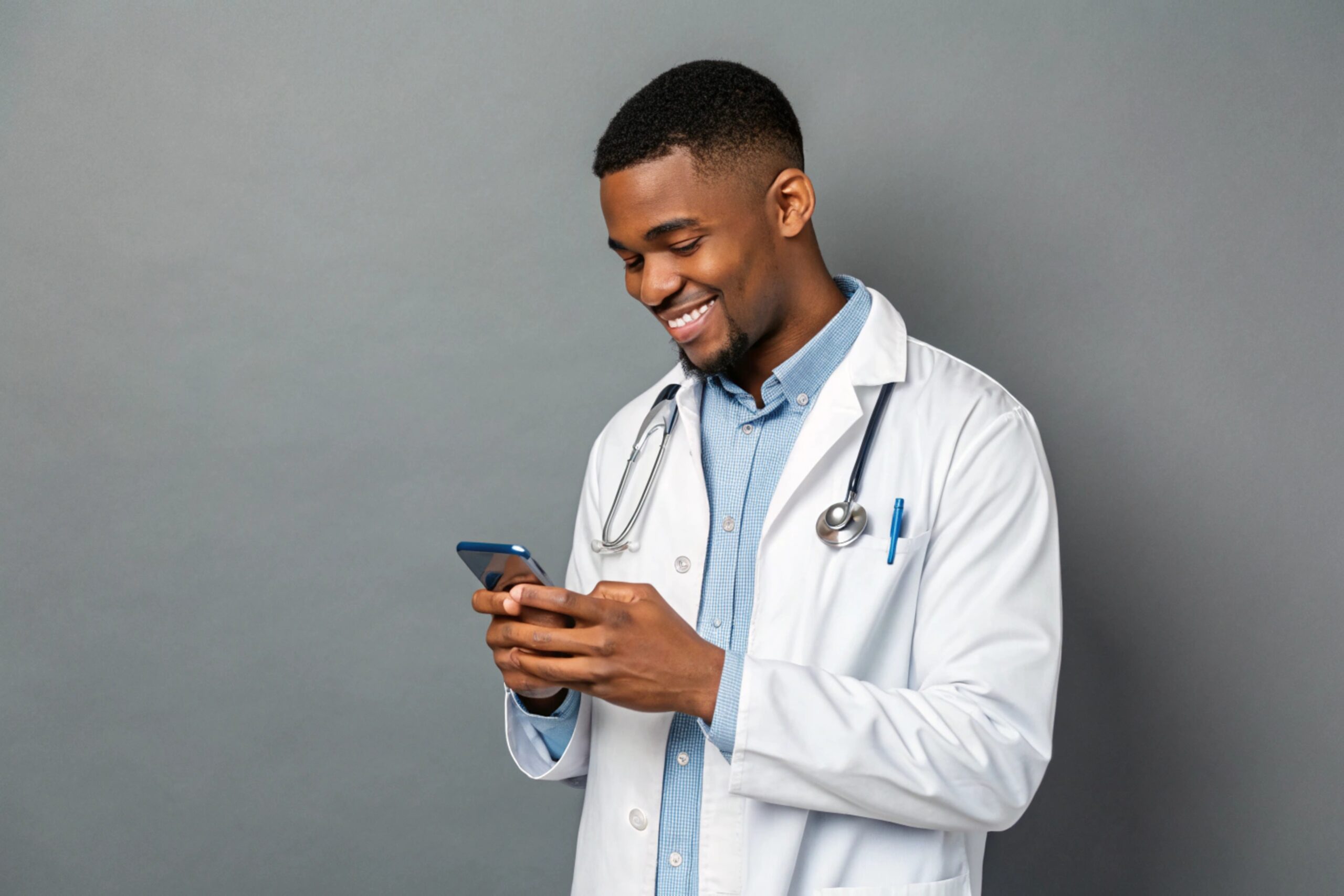 Young male doctor in white coat smiling while looking at screen of his phone, using medical app, standing isolated on gray background